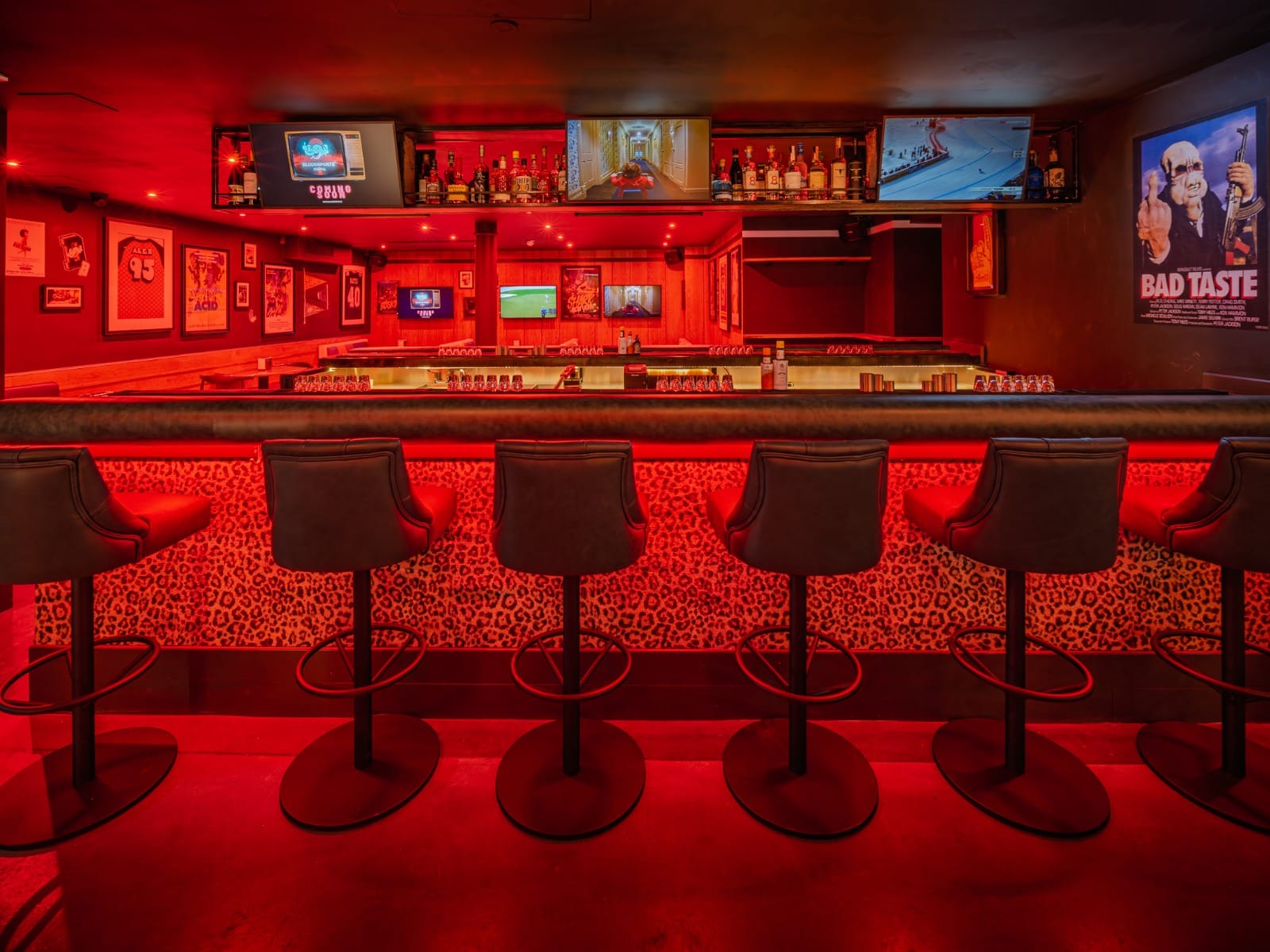 The bar at BLOODsports under deep red light, a row of leather stools along a leopard-print bar front, bottles and glassware racked behind, screens and framed horror posters above.