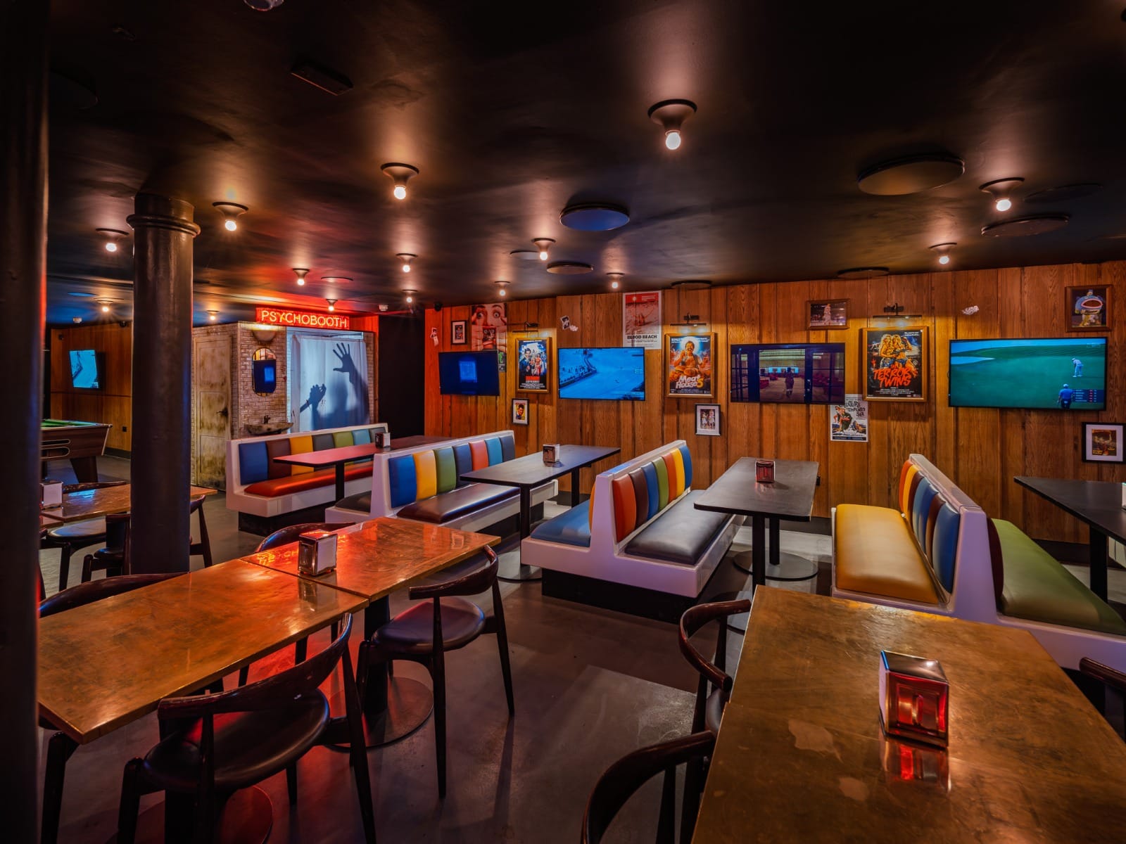 The booth room at BLOODsports: wood-panelled walls hung with horror film posters and screens, striped banquettes in orange, blue and green, copper-topped tables, and a PSYCHOBOOTH sign glowing red beside a shadow of clawing hands.