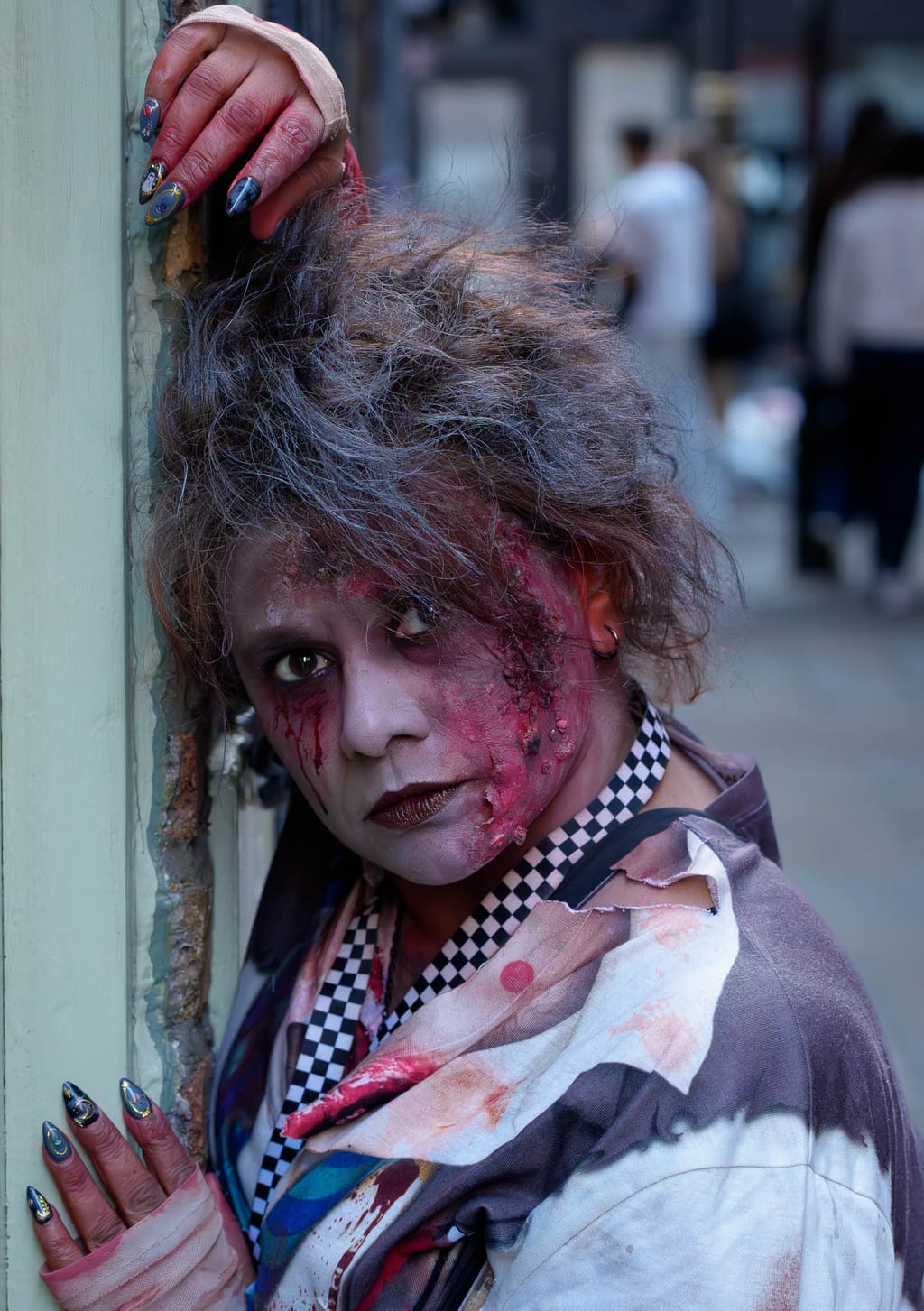 A zombie leans into a painted shopfront doorway, grey-streaked hair backcombed high, a raw prosthetic wound open across her cheek and neck, blood running from one eye, a checkerboard scarf at her throat and a torn printed jacket slipping off one shoulder.