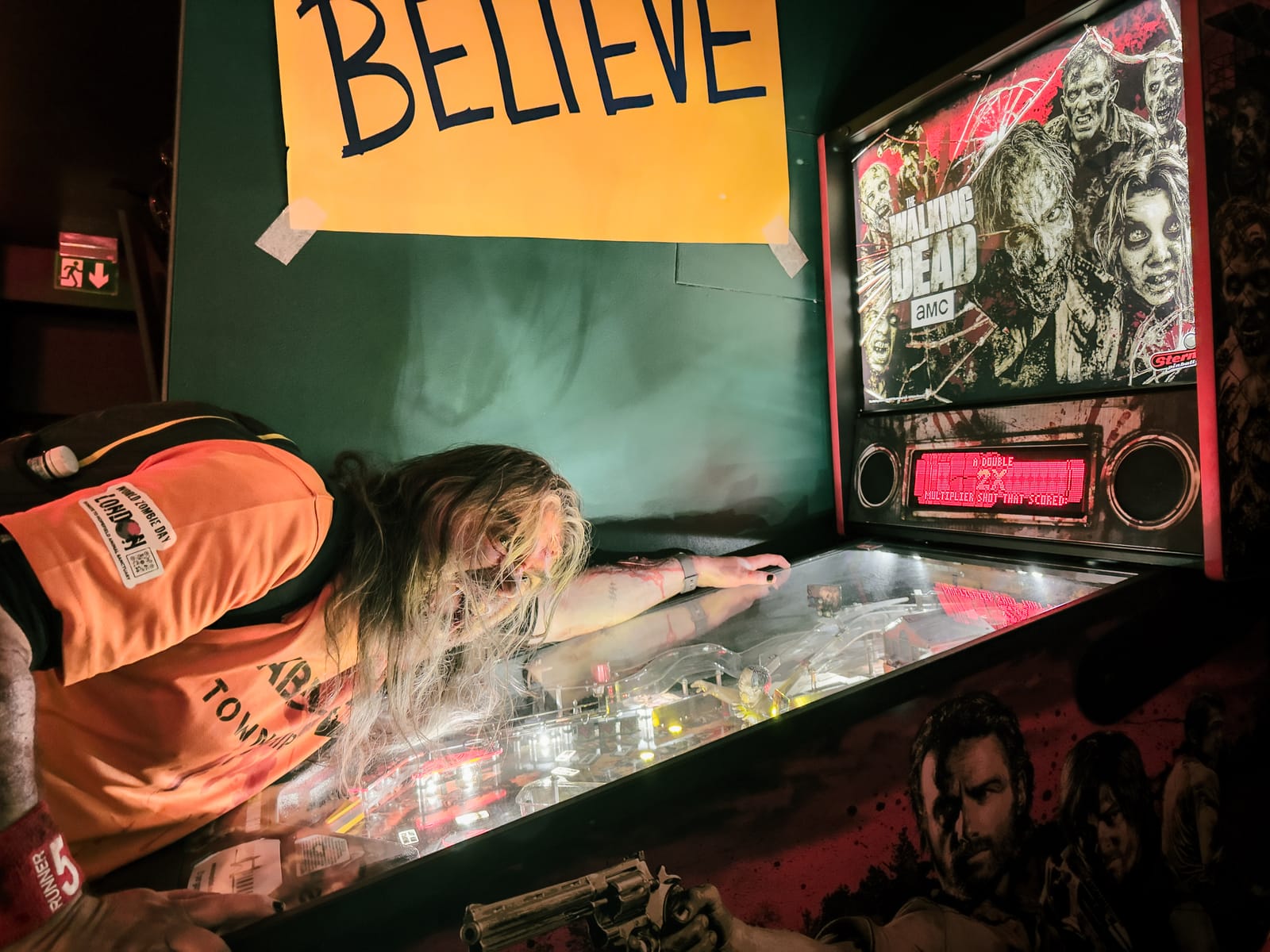 A zombie in a blood-spattered orange World Zombie Day tabard and a Runner 5 armband sprawls over a Walking Dead pinball machine in a dark bar, hair across their face, a hand-lettered BELIEVE sign taped to the wall behind.